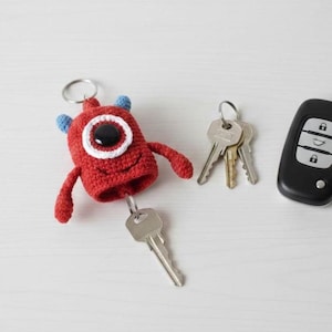 Puede incluir: Un llavero de monstruo rojo tejido a ganchillo con un gran ojo con borde blanco y detalles azules. El llavero sostiene una llave plateada, con un juego de llaves y un mando a distancia negro para el coche cerca. El monstruo tiene brazos pequeños y una boca sonriente.