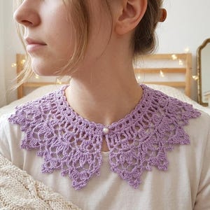 Puede incluir: Un cuello de ganchillo lila con un borde festoneado y un cierre de bot&oacute;n de perla. El cuello se lleva sobre una camisa blanca. El ganchillo tiene un dise&ntilde;o abierto y de encaje.