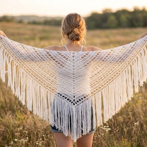 Puede incluir: Un chal de ganchillo blanco con flecos. El chal se sostiene abierto, mostrando el intrincado diseño de tejido abierto. Los flecos cuelgan de la parte inferior y los lados del chal. El chal tiene forma de triángulo.