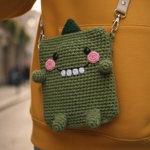 May include: A green crochet crossbody bag shaped like a friendly monster. The bag features a small green fin, pink cheeks, black eyes, and white teeth. It has a beige strap and silver clasps.