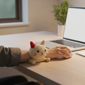 May include: A cream-coloured crocheted cat wrist rest with a red ear. The cat has black stitched details for the eyes, nose, and whiskers. The wrist rest is placed on a person's arm, next to a laptop and mouse on a light-coloured desk.