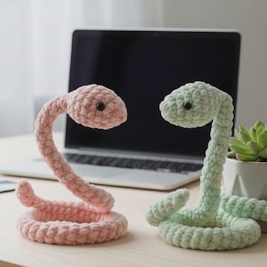 Pode incluir: Dois brinquedos de cobra de crochê. Um é rosa claro, o outro verde claro. Ambos têm olhos de botão pretos. A cobra rosa está enrolada, com a cabeça levantada. A cobra verde também está enrolada, com a cabeça levantada. Um laptop e uma pequena suculenta em vaso estão no fundo.