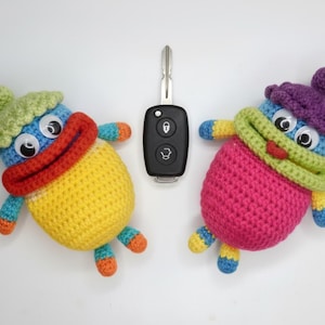 Può includere: Due bambole all'uncinetto fatte a mano con grandi occhi mobili, una gialla e l'altra rosa, affiancano una chiave dell'auto. Le bambole hanno dettagli colorati, tra cui cappelli, sciarpe e arti. Lo sfondo è bianco.