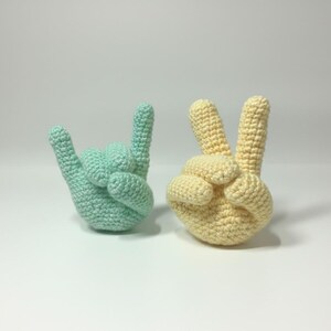 May include: Two crocheted hand sculptures. One is light green, shaped in the "I love you" hand gesture. The other is yellow, forming the peace sign. Both are on a white surface.