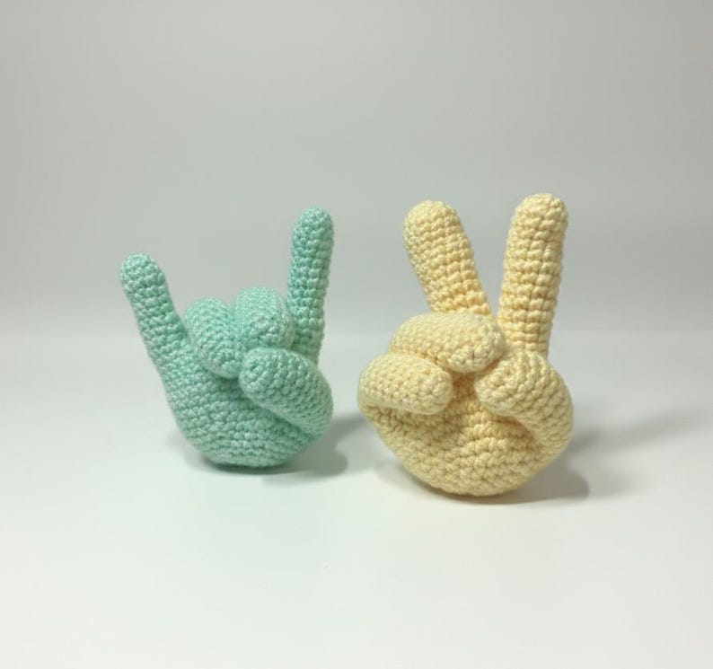 May include: Two crocheted hand sculptures. One is light green, shaped in the "I love you" hand gesture. The other is yellow, forming the peace sign. Both are on a white surface.