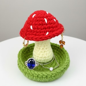 May include: A handmade crochet mushroom-shaped jewelry holder. The red cap has white spots, the stem is cream, and the base is green. A ring, necklace, and earrings are displayed on the holder. The jewelry holder is a unique home decor item.