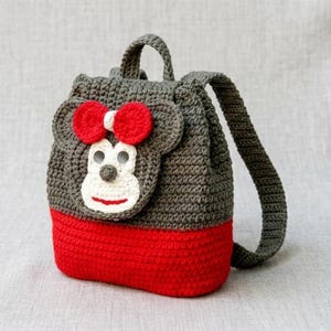 May include: A handmade crochet backpack in gray and red, featuring a monkey face design. The backpack has a red bottom, gray body, and a monkey face with a red bow, white face, and a small gray nose. It has a top handle and adjustable straps.