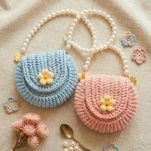 May include: Two crocheted round purses, one light blue and one pink, each with a yellow flower accent. They have pearl-beaded straps and small gold-colored charms. Additional crocheted flowers and a bouquet of dried flowers are scattered around.