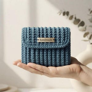 Puede incluir: Una pequeña bolsa rectangular de ganchillo azul con textura acanalada y cierre de solapa. Una etiqueta metálica rectangular está centrada en la solapa con la palabra "handmade" grabada en letras minúsculas negras en la parte frontal.