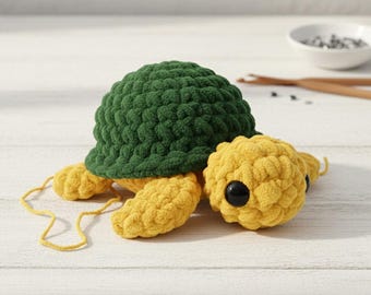 May include: A handmade crochet turtle with a green shell and yellow head, feet, and tail. The turtle has black eyes and is lying on a white surface. The crochet work has a textured, plush appearance.