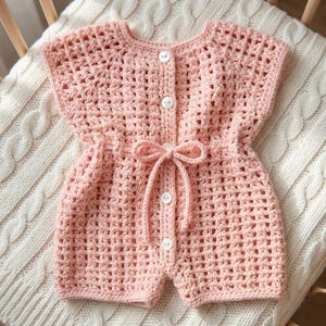 May include: A light pink, crocheted baby romper with short sleeves and shorts. The romper features a button-up front with white buttons and a drawstring bow at the waist. The garment is displayed on a white, textured blanket.