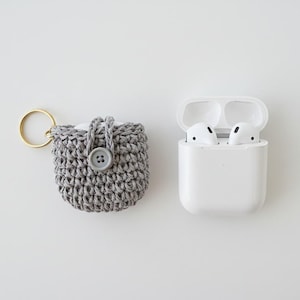Puede incluir: Una funda gris de AirPod tejida a ganchillo con cierre de botón y un llavero dorado junto a una funda de carga blanca de AirPod con dos auriculares blancos en su interior. La funda está hecha a mano y tiene un aspecto texturizado.