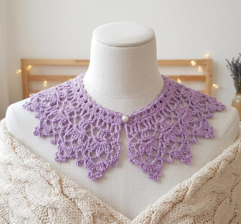 Puede incluir: Un cuello de ganchillo lila con borde festoneado y cierre de bot&oacute;n de perla. El cuello se muestra en un maniqu&iacute; y presenta un delicado patr&oacute;n de tejido abierto. Un accesorio de moda.