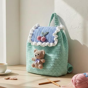 Könnte beinhalten: Handgefertigter gehäkelter Rucksack in Helltürkis. Die Klappe ist hellblau mit weißem Rüschenrand und Kirschdesign. Ein kleiner Teddybär mit lila Schleife ist vorne befestigt. Die Tasche hat einen Griff und einen Kordelzugverschluss.
