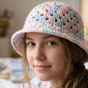 May include: A pastel-colored crochet bucket hat in shades of pink, blue, and cream. The hat features a classic granny square design and a slightly downward-sloping brim. The hat is worn by a person.