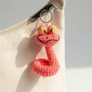 May include: A coral pink crocheted snake keyring featuring a coiled body, a rounded head with large yellow eyes, and a stitched black mouth. The small stuffed creature is attached to a silver metal split ring for easy attachment to bags.