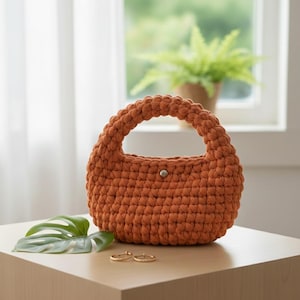 Puede incluir: Un bolso de mano de ganchillo naranja quemado, de forma redondeada y con un asa resistente. El bolso tiene un cierre de botón y se muestra sobre una superficie de color claro. También se ven una hoja verde y dos anillos dorados.