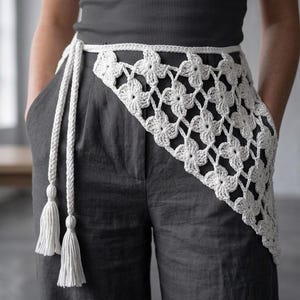 May include: A white crochet belt with a floral pattern and long braided ties with tassels. The belt is worn over gray pants and a gray top. The crochet design is open and airy.