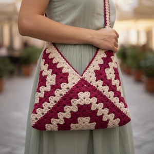May include: A handmade crochet bag in a geometric pattern. The bag features a deep red and cream color scheme, with a matching strap. The bag is held by a person wearing a light green dress. The bag's design is a striking example of crochet craftsmanship.