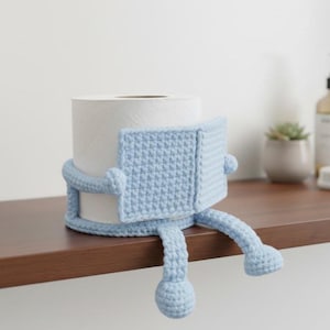 Peut inclure: Un porte-papier toilette en crochet bleu clair en forme de personnage, avec des bras, des jambes et un corps carré. Le support est conçu pour envelopper un rouleau de papier toilette blanc, ajoutant une touche fantaisiste à la décoration de la salle de bain.