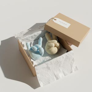 May include: A pair of crochet hand gestures, one light blue and one cream, inside a cardboard box with a tag. The hands are in the 'peace' or 'rock on' position. The box is lined with white tissue paper.