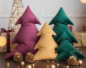 May include: Four plush Christmas tree-shaped pillows in shades of burgundy, gold, and green. The pillows are arranged on a dark surface with gold pinecones, ornaments, and string lights. A decorative gold wire tree is in the background.