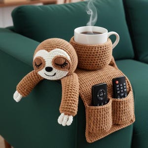 Può includere: Un bracciolo per divano a forma di bradipo all'uncinetto marrone con una tazza di caffè bianca. Il bradipo ha gli occhi chiusi e un viso sorridente. Il bracciolo ha due tasche che contengono telecomandi. La tazza è racchiusa in un supporto all'uncinetto.