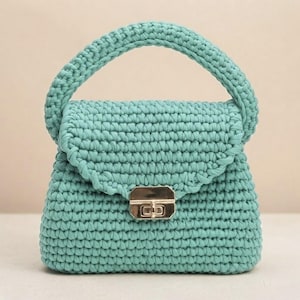 May include: A mint green crochet handbag featuring a thick, rounded top handle and a structured flap closure. The bag is crafted from a dense, textured yarn stitch pattern and is secured at the front with a polished gold-tone metal push-lock clasp.