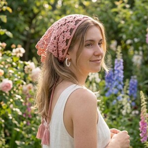 May include: A light pink crocheted headscarf with a floral pattern. The head covering has long tassels and is worn by a person in a garden setting. The headscarf is made of a soft, textured yarn.