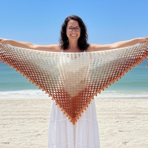 May include: A crocheted shawl with a triangular shape and a gradient color scheme, transitioning from cream to tan and ending in a rust-colored border. The shawl is held up against a backdrop of a beach and blue sky.
