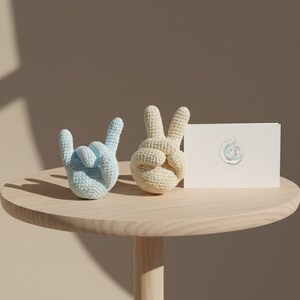 May include: Two crocheted hand sculptures, one light blue and one cream-colored, sit on a wooden table. The blue hand forms the 'rock on' gesture, while the cream hand makes the peace sign. A white card with a circular design is also on the table.