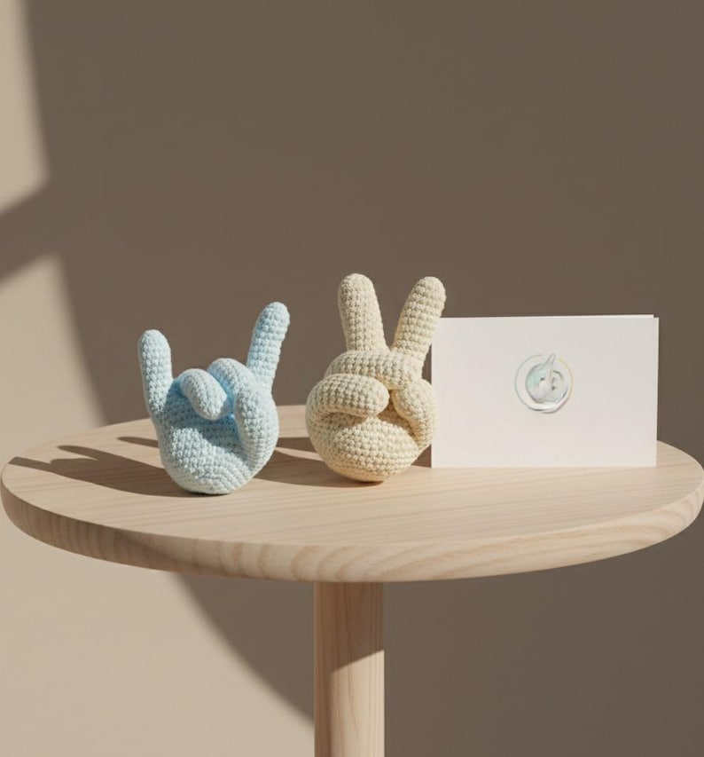 May include: Two crocheted hand sculptures, one light blue and one cream-colored, sit on a wooden table. The blue hand forms the 'rock on' gesture, while the cream hand makes the peace sign. A white card with a circular design is also on the table.