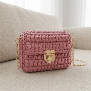 Puede incluir: Un bolso de ganchillo en color rosa empolvado con un tejido de hilo grueso y texturizado y una solapa rectangular. El bolso se asegura con un cierre de presión de metal dorado pulido e incluye una correa de cadena metálica a juego.