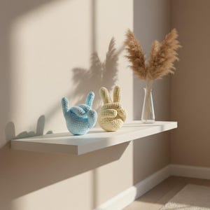 May include: Two crochet hand sculptures, one light blue and one beige, sit on a white shelf. The blue hand is in a rock-on gesture, while the beige hand forms a peace sign. A vase with dried pampas grass is also on the shelf.