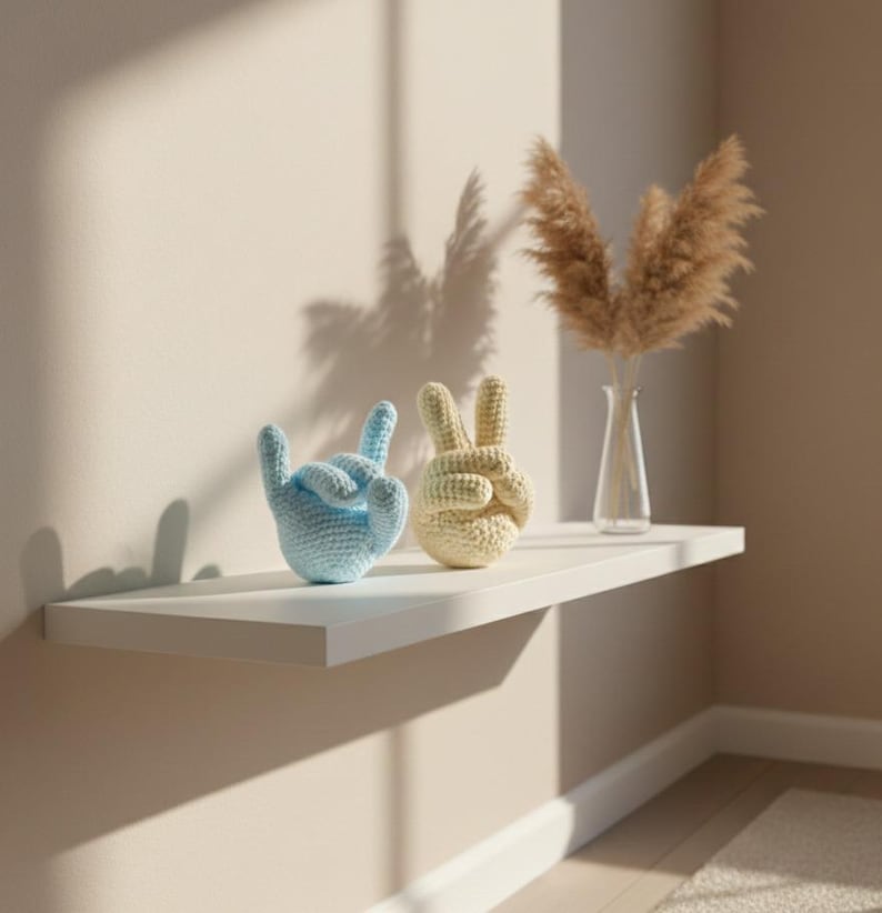 May include: Two crochet hand sculptures, one light blue and one beige, sit on a white shelf. The blue hand is in a rock-on gesture, while the beige hand forms a peace sign. A vase with dried pampas grass is also on the shelf.