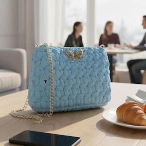 May include: A light blue, crocheted handbag with a gold chain strap and a gold butterfly clasp. The bag is rectangular and sits on a wooden table. A smartphone and a plate with a croissant are also on the table.