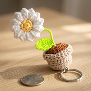 May include: A crocheted flower keychain featuring a white daisy with a yellow center, green stem and leaf, and a brown pot. The keychain is attached to a silver metal ring. The flower is approximately 2 inches tall.