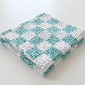 Puede incluir: Una manta cuadrada doblada que presenta un patrón de cuadros repetitivo en colores verde menta y blanco alternos. El tejido tiene una construcción visible de punto de ganchillo texturizado, creando un diseño de cuadrícula uniforme en toda la superficie.