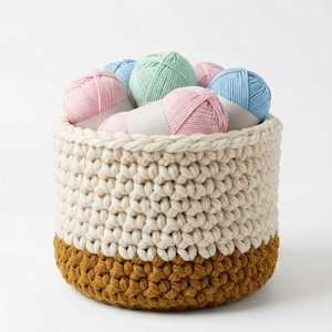 May include: A round, cream-colored crochet basket with a mustard-colored base, filled with pastel-colored yarn balls. The yarn colors include pink, blue, and green. The basket is made of thick, textured yarn.
