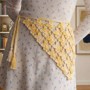 May include: A yellow and cream-colored crochet belt with a floral pattern. The belt is tied at the waist with tassels hanging down. The belt is worn over a floral patterned dress.