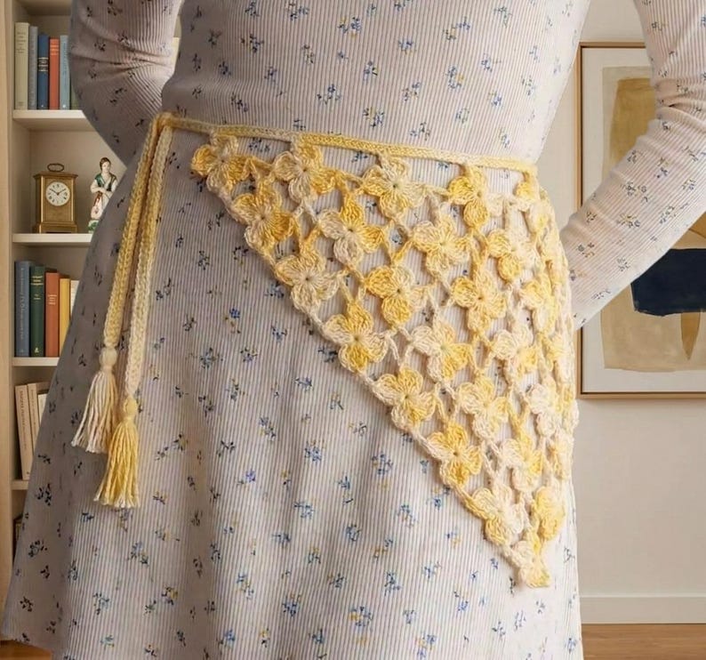 May include: A yellow and cream-colored crochet belt with a floral pattern. The belt is tied at the waist with tassels hanging down. The belt is worn over a floral patterned dress.
