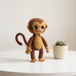 Op de afbeelding: Een gehaakt apenknuffeltje van donkerbruin en beige garen, met grote zwarte plastic ogen en een lange gekrulde staart. De aap heeft een rond hoofd, kleine oren en een lichtbeige borstvlek. Naast de aap staat een vetplantje in een beige pot.
