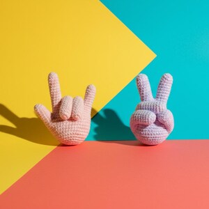 May include: Two crocheted hand sculptures, one in a light pink color forming the 'rock on' hand gesture, and the other in a lavender color forming the peace sign. The background is divided into sections of yellow, teal, and coral.
