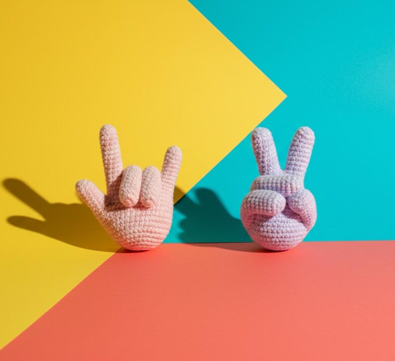 May include: Two crocheted hand sculptures, one in a light pink color forming the 'rock on' hand gesture, and the other in a lavender color forming the peace sign. The background is divided into sections of yellow, teal, and coral.