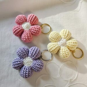 Könnte beinhalten: Drei gehäkelte Blumen-Schlüsselanhänger in Rosa, Gelb und Lila. Jede Blume hat eine weiße Mitte und einen goldenen oder silbernen Schlüsselring. Die Blumen sind aus strukturiertem Garn gefertigt und auf einer weißen Oberfläche angeordnet.