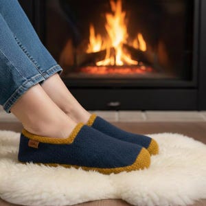 以下が含まれることがあります： マスタードイエローのトリムが付いたネイビーブルーのスリッパが、白いシープスキンのラグの上に置かれています。スリッパはクラシックなデザインです。燃える火のある暖炉が背景にあります。