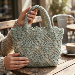 Può includere: Una borsa tote all'uncinetto verde salvia con un bordo superiore smerlato e manici robusti. Il design traforato rivela una fodera di colore neutro. La borsa è tenuta su un tavolo di legno, suggerendo un oggetto fatto a mano.