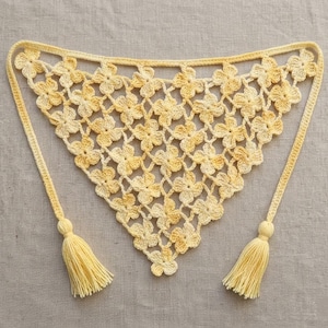 May include: A handmade, yellow crochet shawl with a floral pattern. The shawl has two long ties, each ending in a tassel. The crochet work is detailed, and the color is a soft, warm yellow.