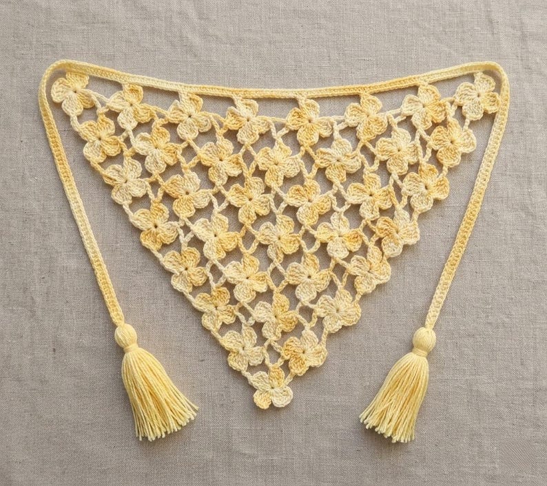 May include: A handmade, yellow crochet shawl with a floral pattern. The shawl has two long ties, each ending in a tassel. The crochet work is detailed, and the color is a soft, warm yellow.