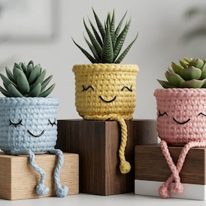 Peut inclure: Trois pots de fleurs en crochet bleus, jaunes et roses, chacun avec un visage souriant et des jambes pendantes. Les pots contiennent des plantes succulentes. Le pot jaune est sur un bloc de bois foncé, tandis que les pots bleus et roses sont sur des blocs de bois plus clairs.